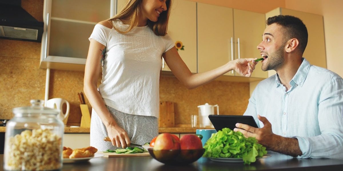 Attractive couple chatting in the kitchen early morning. Beautiful woman feed her husband while cooking breakfast at home